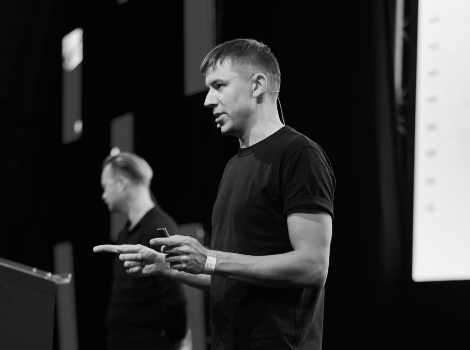 Black and white photo of a Studio 28K founder speaking on stage with a clicker in hand, with his co-founder in the background.