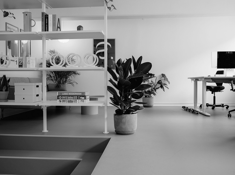 Black and white photo of the Studio 28K office in Copenhagen, with design books, award trophies and plants on white shelves next to a desk.