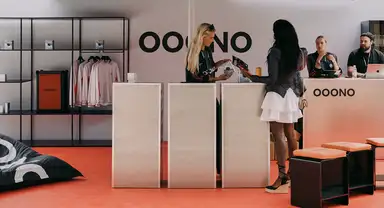 A photo of an event or trade-show booth: two women interact across a reception counter beneath "OOONO" signage, with a clothing rack, an orange product box, and two seated staff members behind the counter, all set on an orange floor.