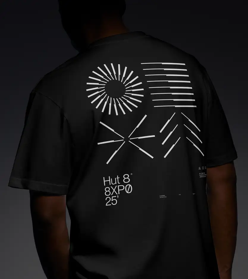 A close-up back view of a person wearing an oversized black Hut 8 branded T-shirt, photographed against a dark moody gray background with dramatic lighting. The shirt's back carries a large white graphic print composed of abstract line-based motifs — a radiating sunburst, horizontal dashed bars, an X-shaped crossing of lines, and a chevron/arrow pattern — all rendered in the Hut 8 visual style. Below the graphics, white text reads "Hut 8® 8XPO 25'" with small technical print, care symbols, a material composition ("51% Polyurethane, 49% Polyamide"), an article code, and the faint tagline "POWERING THE NEXT."