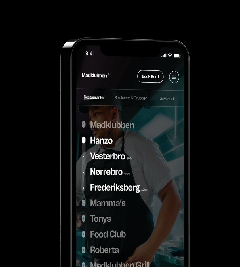 Mockup of a smartphone displaying the Madklubben restaurant app on a black background. The screen shows a dark header with the Madklubben logo, a 'Book Bord' button, and a menu icon, below tabs for Restauranter, Selskaber & Grupper, and Gavekort. A list of restaurant brands is layered over a background photo of a chef in an apron, showing numbered categories (Madklubben with 4 locations, Hanzo with 3, Mamma's, Tonys, Food Club, and Roberta each with 1) and individual Hanzo locations in Vesterbro, Nørrebro, and Frederiksberg with their distances.