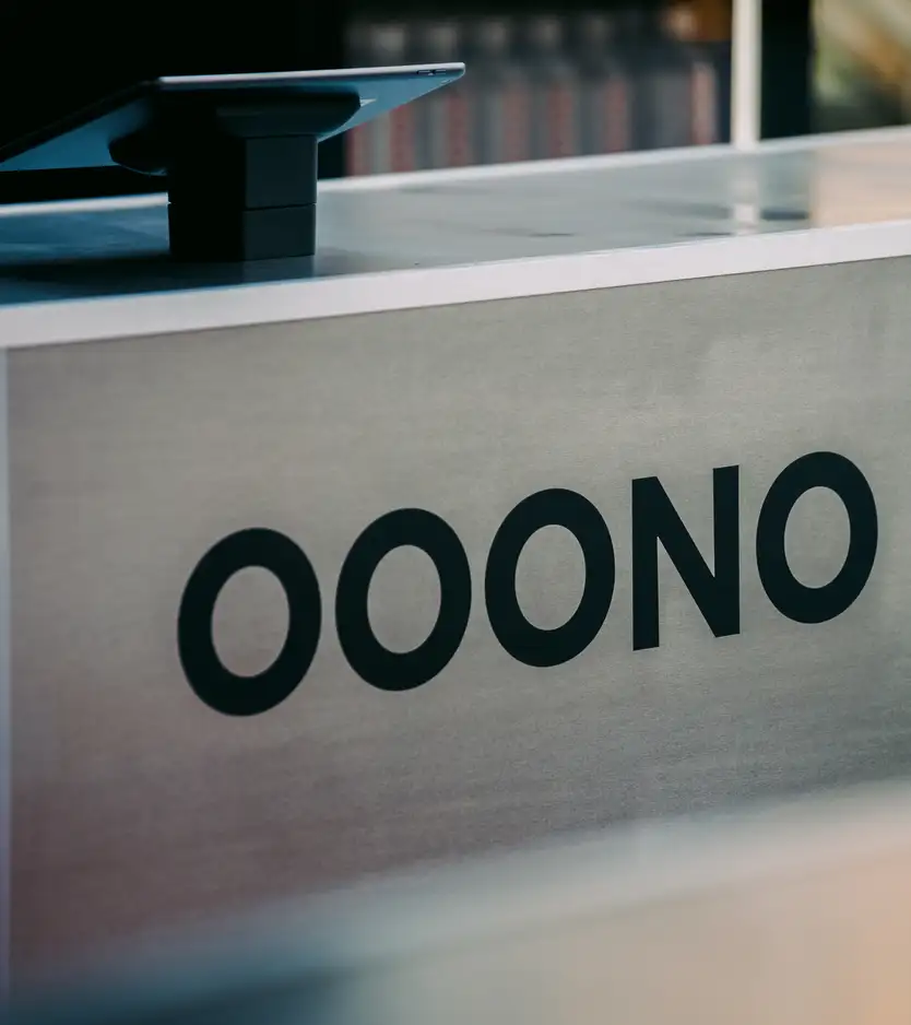 A close-up of a metal reception desk or counter with the black "OOONO" wordmark printed on its front panel, an iPad on a stand visible at the top left, and blurred bottles in the background.