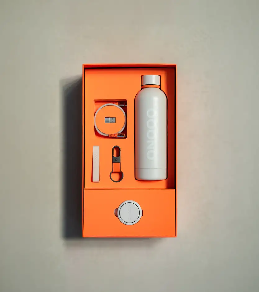 An open orange OOONO gift box containing a water bottle, USB-C cable, keychain, small bar item, and a circular device, all in white/gray tones.