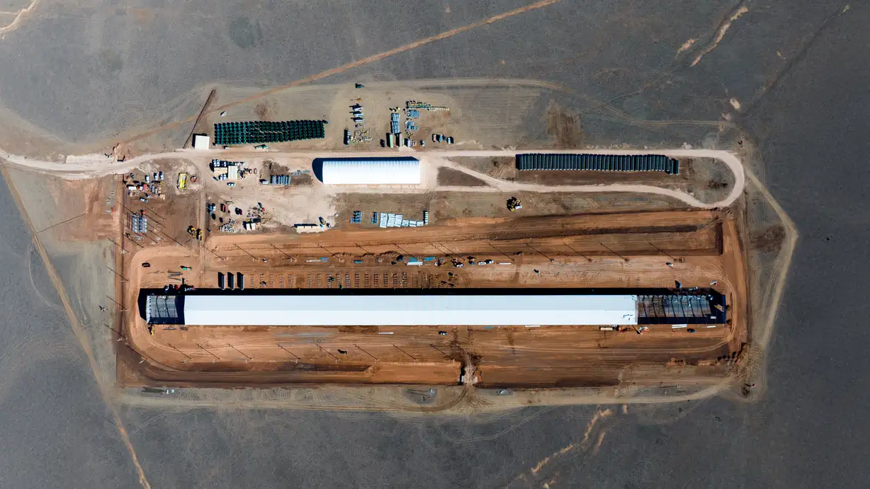 Aerial top-down view of a construction site showing a long warehouse building under construction on a cleared reddish-brown pad, surrounded by staging areas with equipment, materials, and an access road, set in open gray terrain.