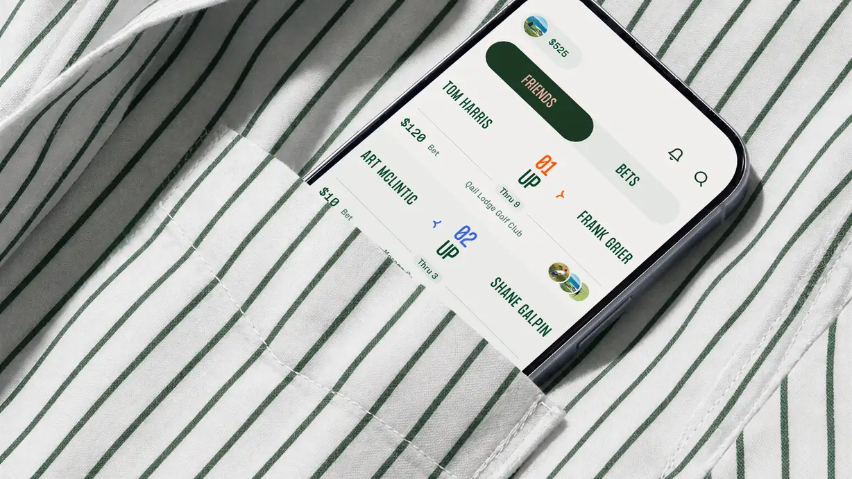 A smartphone tucked into the chest pocket of a white shirt with thin green pinstripes, shown at an angle. The screen displays the Calcutta golf-betting app in the brand's green-and-cream palette. At the top, a user balance reads "$525," with a toggle between "FRIENDS" and "BETS" tabs, plus notification and search icons. Below are head-to-head match bets between friends: "TOM HARRIS" vs. "FRANK GRIER" with a "$120 Bet," showing "1 UP Thru 9" at Quail Lodge Golf Club (orange highlight); and "ART MCLINTIC" vs. "SHANE GALPIN" with a "$10 Bet," showing "2 UP Thru 3" (blue highlight). Small circular profile photos of golfers accompany the entries.