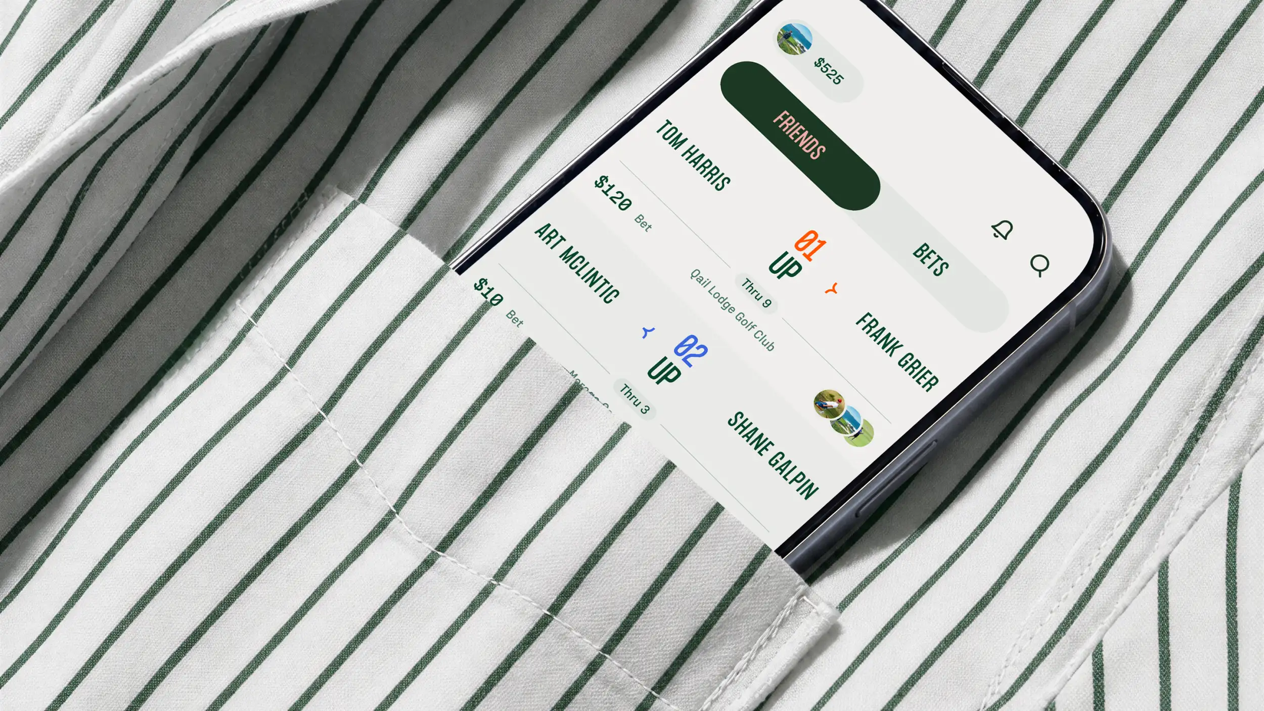 A smartphone tucked into the chest pocket of a white shirt with thin green pinstripes, shown at an angle. The screen displays the Calcutta golf-betting app in the brand's green-and-cream palette. At the top, a user balance reads "$525," with a toggle between "FRIENDS" and "BETS" tabs, plus notification and search icons. Below are head-to-head match bets between friends: "TOM HARRIS" vs. "FRANK GRIER" with a "$120 Bet," showing "1 UP Thru 9" at Quail Lodge Golf Club (orange highlight); and "ART MCLINTIC" vs. "SHANE GALPIN" with a "$10 Bet," showing "2 UP Thru 3" (blue highlight). Small circular profile photos of golfers accompany the entries.