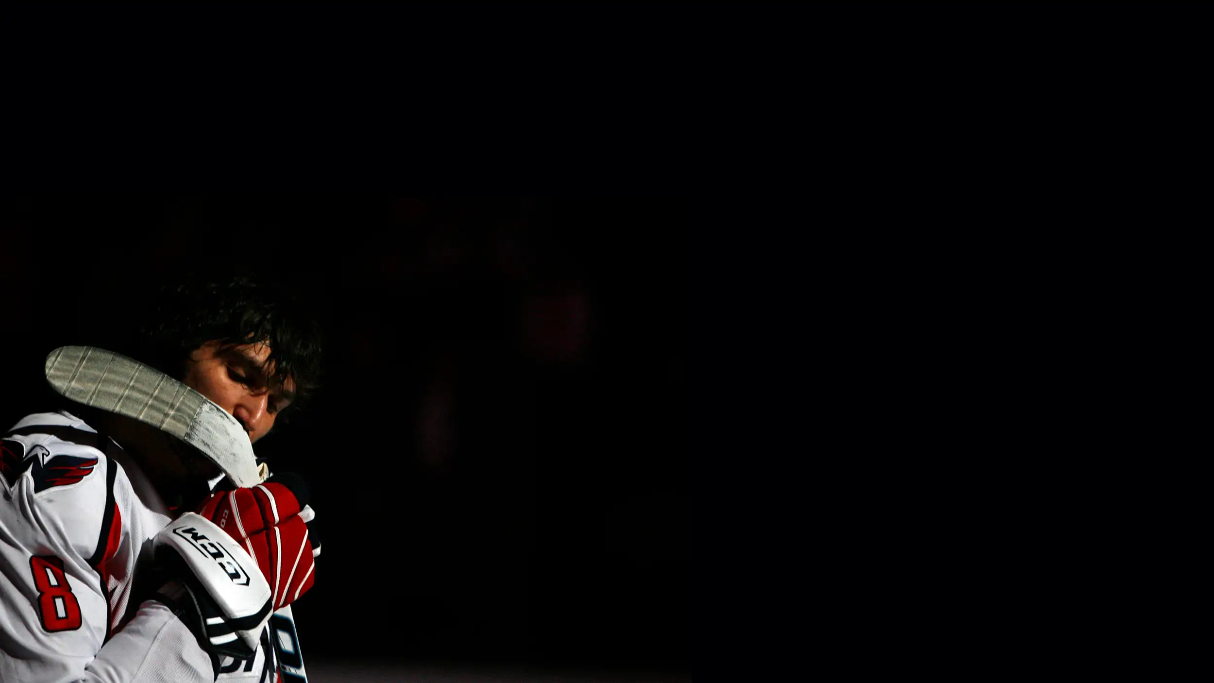 A dramatic, dark, spotlit photo of a hockey player in a moment of quiet focus, positioned on the left side of the frame against a nearly all-black background. He wears a white Washington Capitals jersey with the number 8 and the team's eagle logo, along with red-and-white CCM gloves. His head is bowed and eyes closed as he rests his stick against his shoulder and holds it close, appearing contemplative during what looks like a pre-game moment. The right two-thirds of the image is deep black negative space.