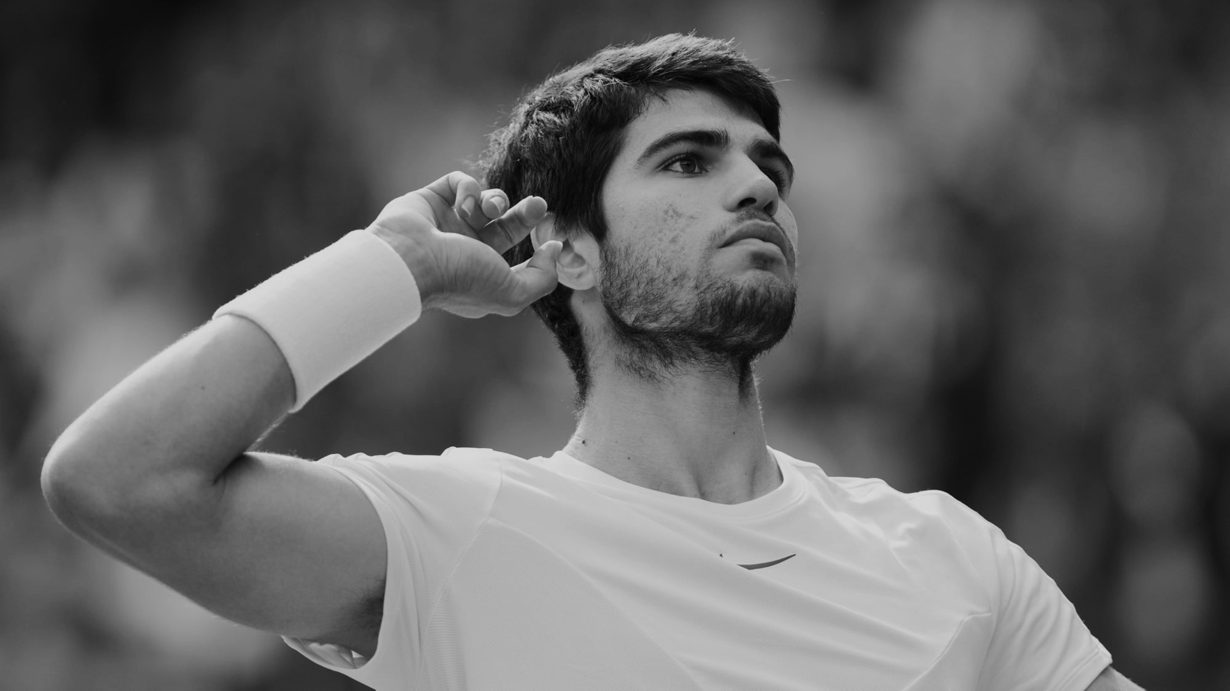 A black-and-white close-up photo of a young male tennis player during a match, shown from the chest up against a softly blurred crowd of spectators. He has dark hair and a short beard, wears a white athletic shirt with a small logo on the chest and a white wristband, and raises one hand to cup his ear toward the crowd in a gesture inviting the audience to cheer. His expression is focused and intense as he looks off to the side.