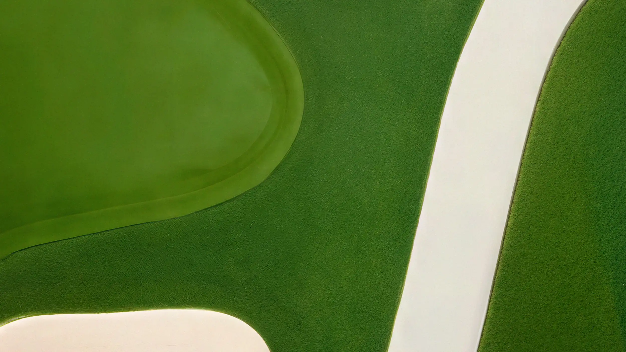 An aerial photo of a golf course green, with bold curved shapes in two shades of green turf separated by cream-colored bunkers or paths, in a minimalist, abstract composition.