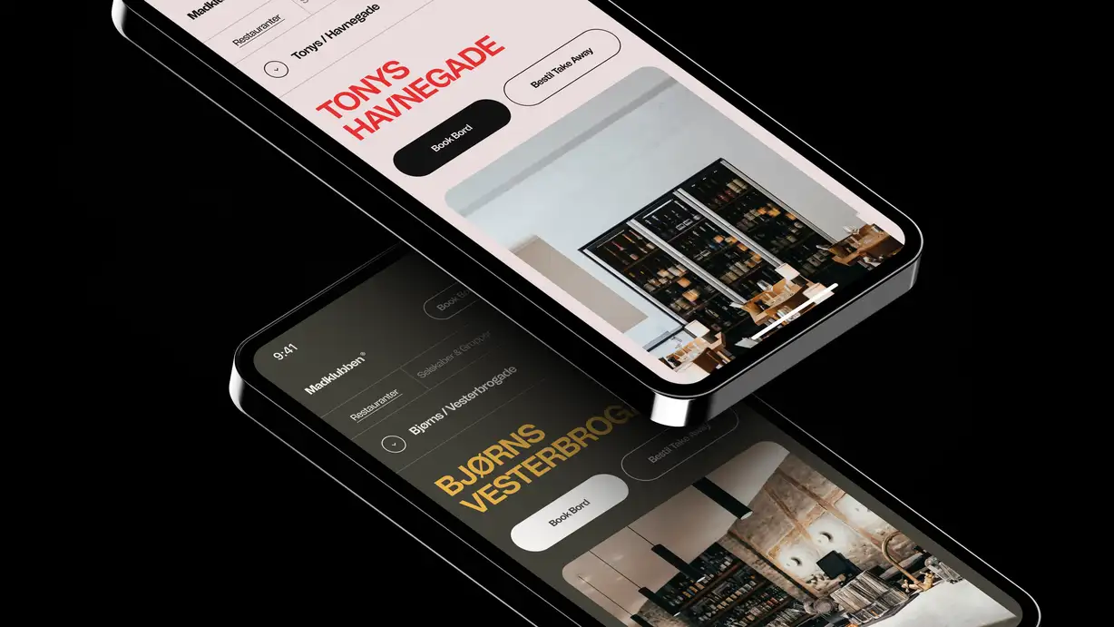 Two overlapping smartphone mockups on a black background, tilted at angles, each showing a Madklubben restaurant page. The upper phone displays 'Tonys Havnegade' in bold red type on a light pink background, with 'Book Bord' and 'Bestil Take Away' buttons above a photo of a dimly lit restaurant with wine shelves and wood furniture. The lower phone displays 'Bjørns Vesterbrogade' in bold gold type on a dark background, with matching 'Book Bord' and 'Bestil Take Away' buttons above a photo of an industrial-style space with exposed pipes, brick walls, and dark cabinetry."  Shorter version: "Two tilted smartphone mockups showing Madklubben restaurant pages for Tonys Havnegade (light theme, red text) and Bjørns Vesterbrogade (dark theme, gold text), each with Book Bord and Take Away buttons over interior photos.