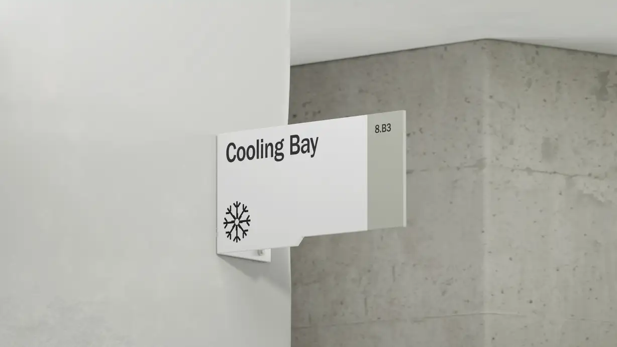 A minimalist white wayfinding sign mounted so it projects from the corner of a white wall, next to an exposed gray concrete wall. The flag-style sign reads "Cooling Bay" in bold black type, with the room code "8.B3" on a light gray band to the right and a black snowflake icon in the lower-left corner. The clean, modern signage suits an industrial facility interior.