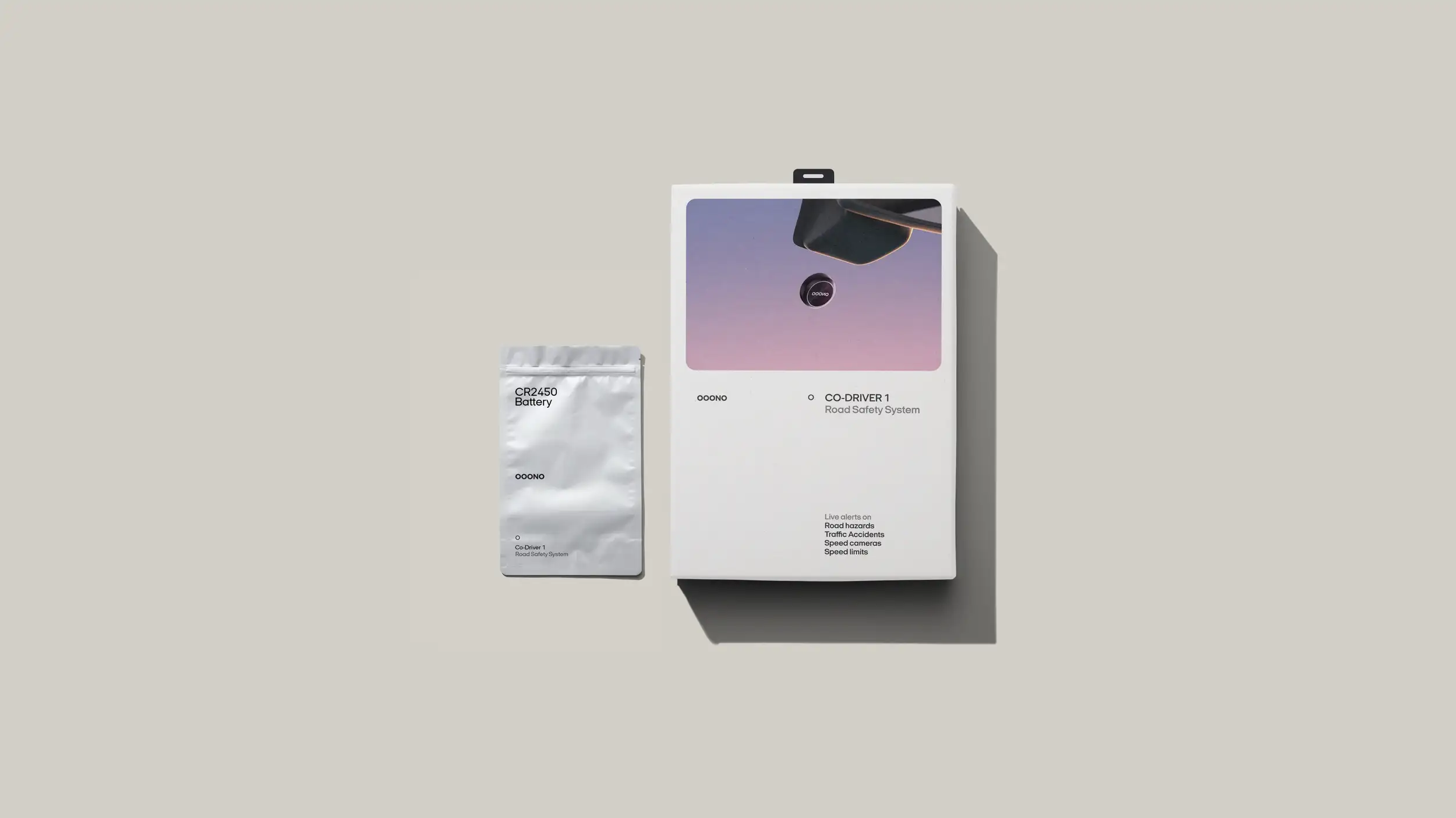 A flat-lay of OOONO Co-Driver 1 product packaging on a warm gray background, casting a soft shadow. On the right is the main white retail box with a hanging tab at the top; its upper half shows a purple-to-pink gradient photo of the black OOONO device mounted on a car dashboard. Below, the box is labeled with the "OOONO" wordmark, a small circular "O" icon, and "CO-DRIVER 1 — Road Safety System," with a feature list in the lower corner reading "Live alerts on: Road hazards, Traffic Accidents, Speed cameras, Speed limits." To the left sits a smaller silver foil pouch labeled "CR2450 Battery," also branded with the "OOONO" wordmark and "Co-Driver 1 — Road Safety System."