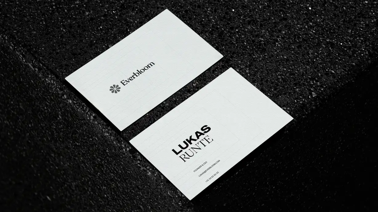 Two white business cards resting on a dark, textured black stone or terrazzo surface with dramatic, moody lighting. The upper card shows the front: the "Everbloom" wordmark beside a small floral/starburst logo, centered on the white card. The lower card shows the back: the name "LUKAS RUNTE" in bold black type, with the title "FOUNDER & CEO," the email "LUKAS@EVERBLOOM.COM," and a phone number "+31 24 02 62 42" beneath it. The clean, minimalist branding contrasts sharply with the speckled dark background.