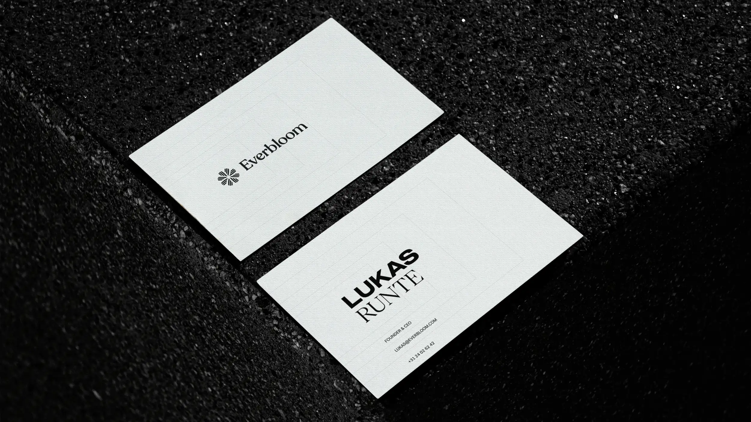 Two white business cards resting on a dark, textured black stone or terrazzo surface with dramatic, moody lighting. The upper card shows the front: the "Everbloom" wordmark beside a small floral/starburst logo, centered on the white card. The lower card shows the back: the name "LUKAS RUNTE" in bold black type, with the title "FOUNDER & CEO," the email "LUKAS@EVERBLOOM.COM," and a phone number "+31 24 02 62 42" beneath it. The clean, minimalist branding contrasts sharply with the speckled dark background.