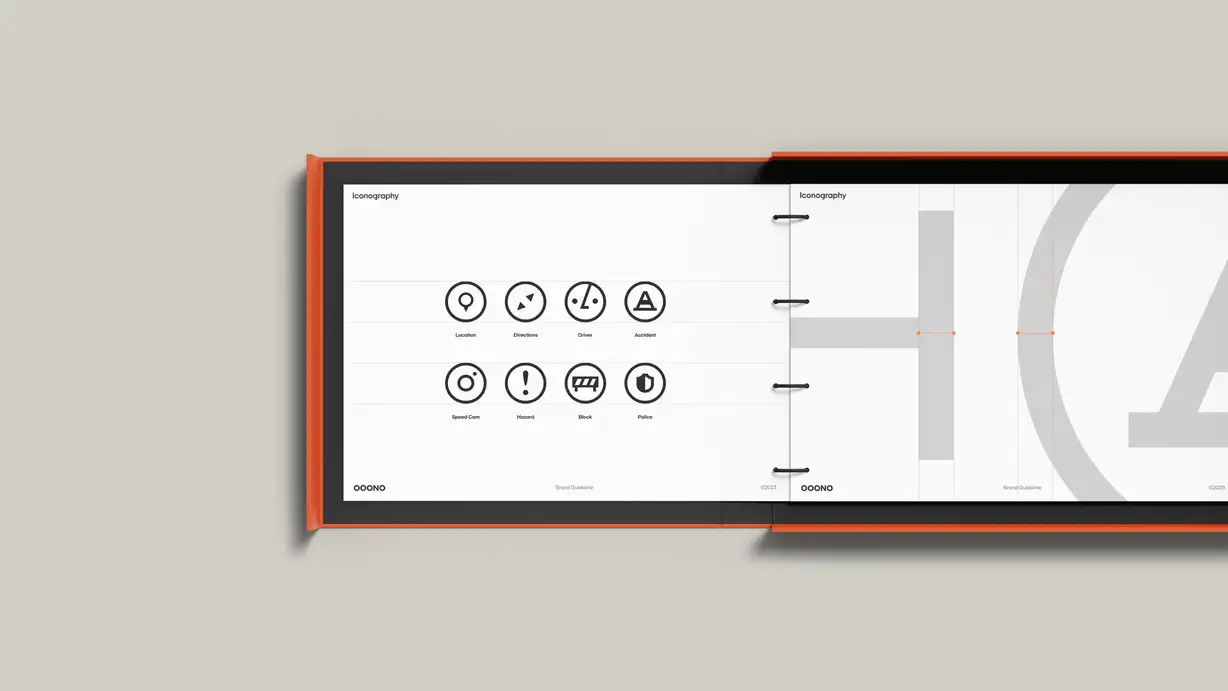 A wide, landscape-format OOONO brand guideline book lying open on a warm gray surface, bound with an orange cover and black ring binding down the center. Both visible pages are headed "Iconography." The left page displays eight black circular line icons in a two-row grid, each labeled: Location (map pin), Directions (arrow), Driver, and Accident (warning triangle) on the top row; Speed Cam, Hazard (exclamation mark), Block (barrier), and Police (shield) on the bottom row. The right page shows an enlarged construction view of an icon with light gray shapes and orange grid guidelines demonstrating how the icons are built. Both pages carry the "OOONO" wordmark, the footer "Brand Guideline," and a "©2023" mark.