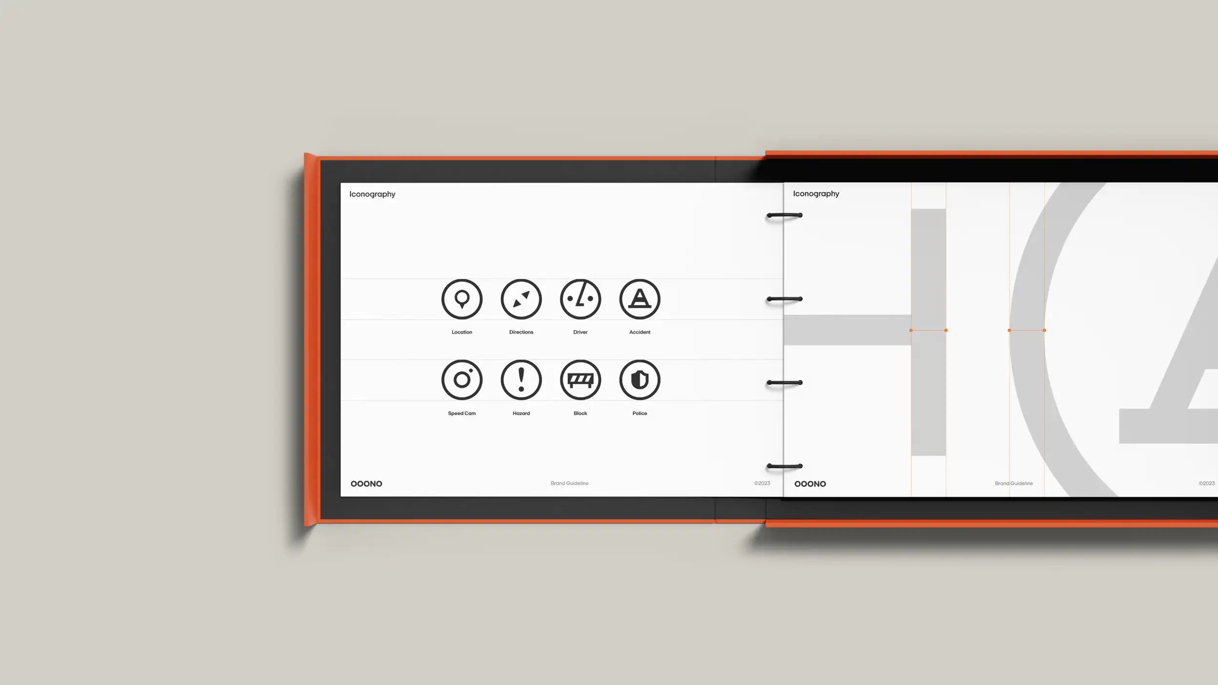 A wide, landscape-format OOONO brand guideline book lying open on a warm gray surface, bound with an orange cover and black ring binding down the center. Both visible pages are headed "Iconography." The left page displays eight black circular line icons in a two-row grid, each labeled: Location (map pin), Directions (arrow), Driver, and Accident (warning triangle) on the top row; Speed Cam, Hazard (exclamation mark), Block (barrier), and Police (shield) on the bottom row. The right page shows an enlarged construction view of an icon with light gray shapes and orange grid guidelines demonstrating how the icons are built. Both pages carry the "OOONO" wordmark, the footer "Brand Guideline," and a "©2023" mark.