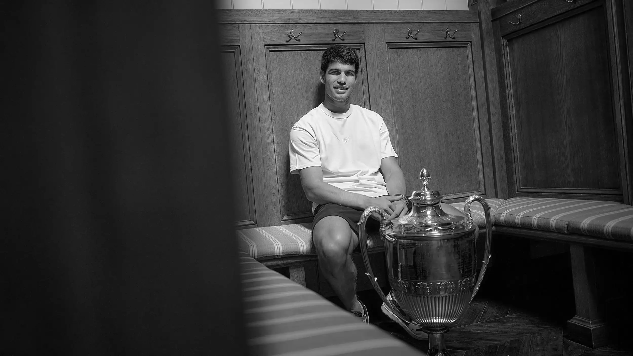 A black-and-white photo of a young male tennis player seated on a striped bench in an elegant, wood-paneled locker room with coat hooks along the walls. He wears a plain white T-shirt, shorts, and sneakers, smiling warmly toward the camera with a relaxed, contented posture. In the foreground on the right stands a large, ornate silver trophy cup with two handles and a lidded, fluted design, one of his hands resting near it. The left portion of the frame is in deep shadow, and part of a bench extends across the bottom foreground.