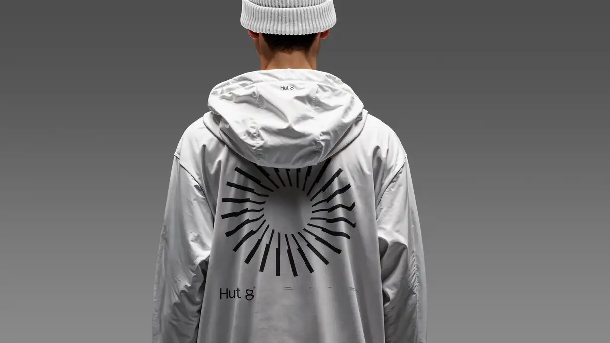 Rear view of a person wearing a white beanie and an oversized white hoodie printed with a black sunburst graphic and "Hut 8" branding, against a gray studio background.