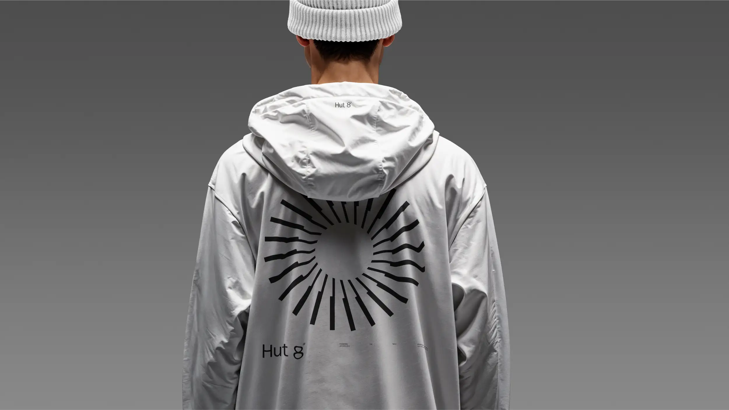 Rear view of a person wearing a white beanie and an oversized white hoodie printed with a black sunburst graphic and "Hut 8" branding, against a gray studio background.