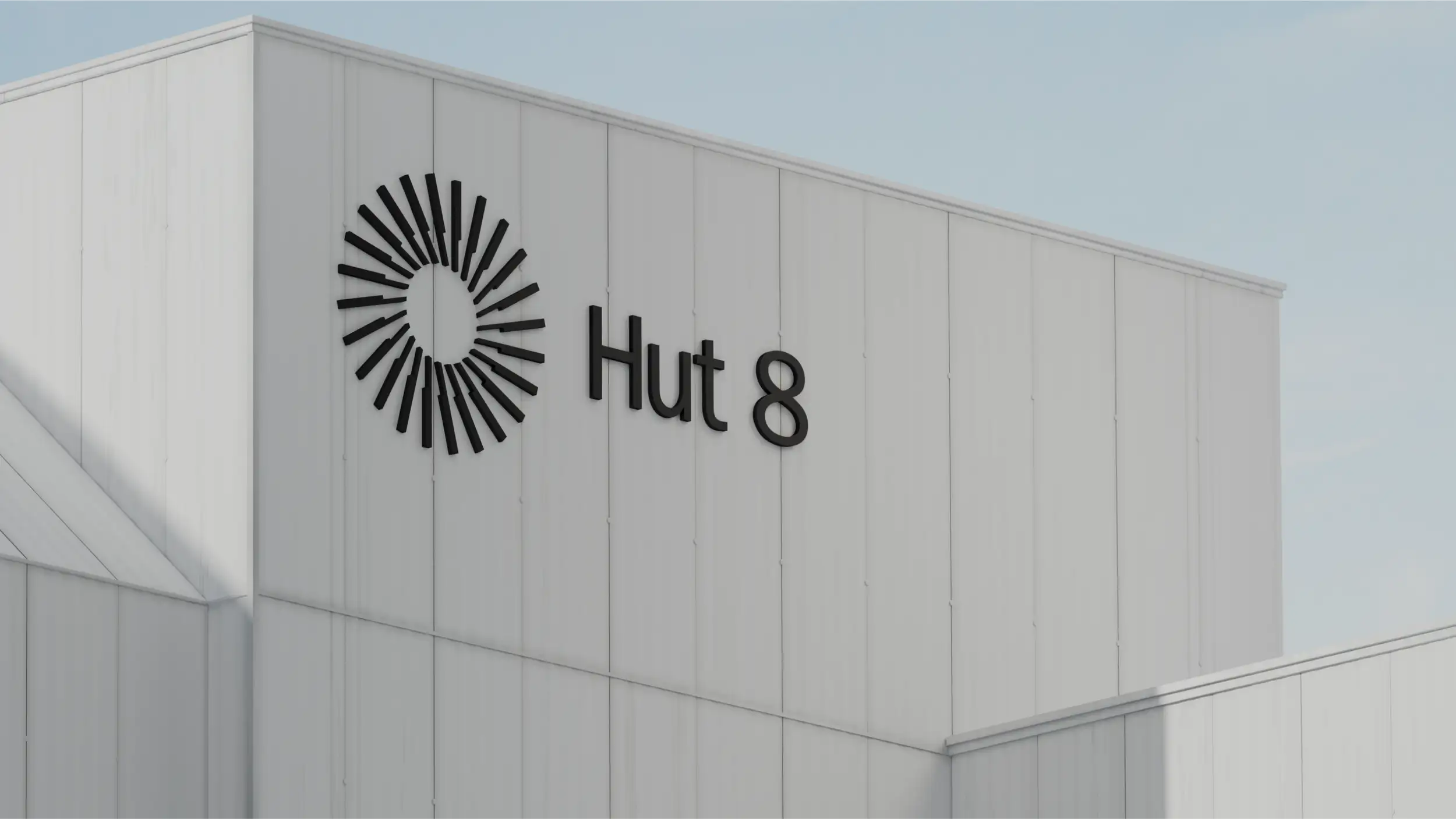 A low-angle exterior shot of a clean, white corrugated-metal industrial building against a clear pale blue sky. Mounted on the upper facade is the "Hut 8" brand signage in black: a circular sunburst/pinwheel logo made of radiating tapered lines beside the wordmark "Hut 8." Soft daylight and gentle shadows give the minimalist, modern structure a crisp, corporate look.