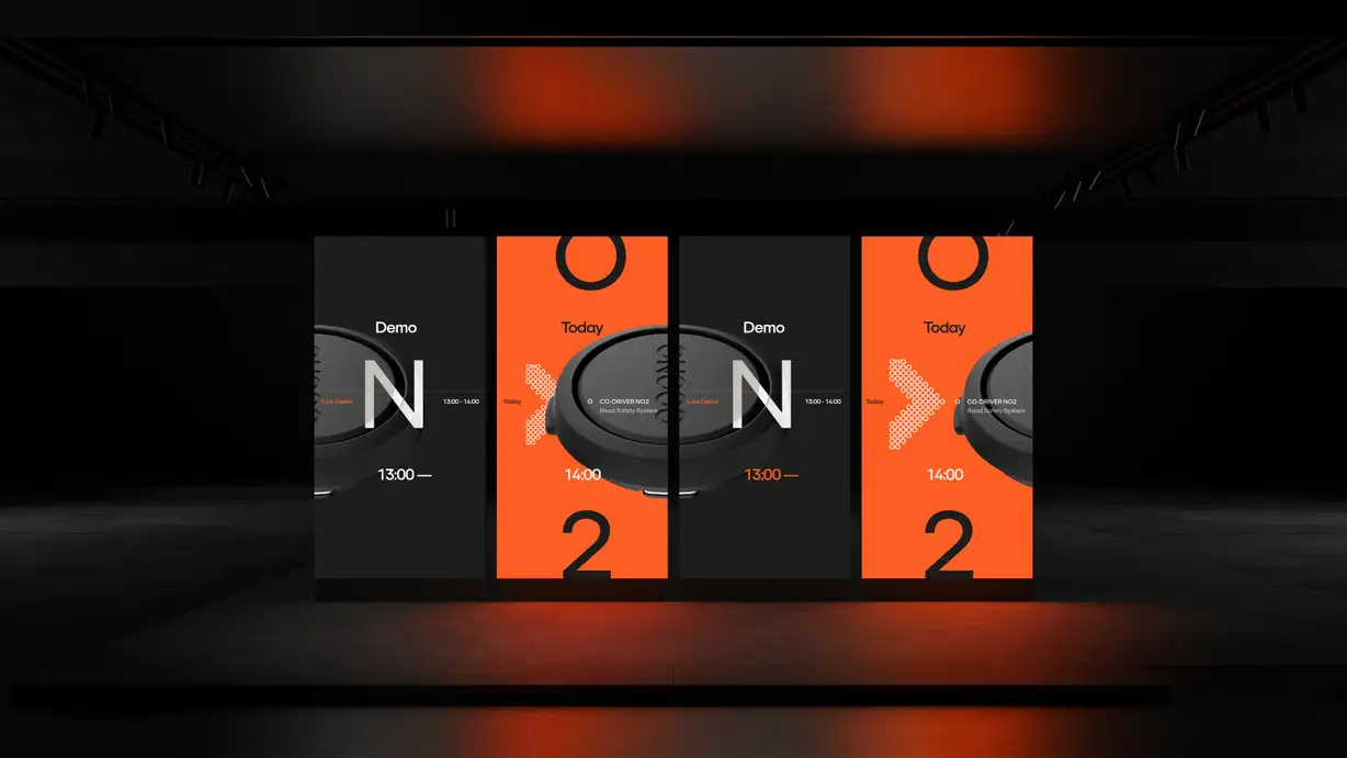 A dark showroom installation with four tall vertical LED panels alternating black and orange, displaying a black circular device alongside text like "Demo," "Today," "CO-DRIVER NO2 Road Safety System," time stamps "13:00–14:00," and large numerals, under warm orange ambient lighting.