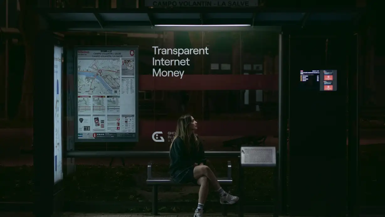A moody nighttime photo of a young woman sitting alone on a bench inside a lit bus shelter, waiting. She has long hair and wears a denim jacket, shorts, white socks, and sneakers, gazing off to the side. The shelter is at the "Campo Volantín – La Salve" stop (Bilbobus), with a route map and timetable panel on the left and a digital arrivals display on the right. A large glass GHO advertisement behind her reads "Transparent Internet Money" in white, with the "G" face logo and "GHO. By Aave ↓ 5..." branding partly obscured by her figure. Dark trees and city buildings are faintly visible beyond the glass.