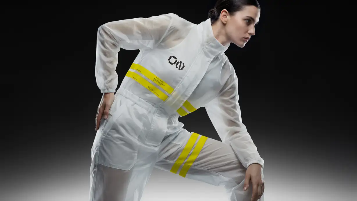A woman in a sheer white technical jumpsuit with yellow reflective stripes and the ON logo on the chest, caught mid-motion in an athletic lunging pose against a black background.