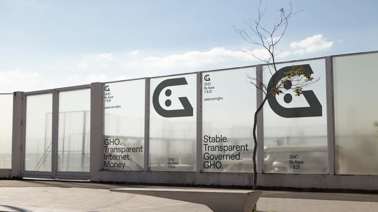 An outdoor urban scene showing a row of frosted-glass fence panels displaying large GHO advertising posters, under a bright blue sky with scattered clouds. A concrete bench and a slender bare-branched sapling stand in the paved foreground. The posters carry the GHO branding: one reads "GHO. Transparent Internet Money" with the "G" face logo and "GHO. By Aave ↑ 8.15 / aave.com/gho"; the center panels show an enlarged black "G" face logo; and another reads "Stable. Transparent. Governed. GHO." with "GHO. By Aave ↑ 8.21." The frosted panels are semi-transparent, revealing industrial structures and fencing behind them.