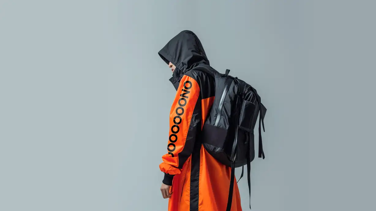 A side-profile photo of a person in a black hood and an orange-and-black colorblocked long coat with a black backpack, "OOONO" printed vertically down the orange sleeve, against a plain gray background.