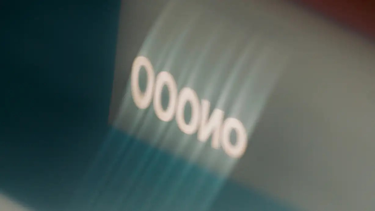 A close-up, motion-blurred shot of the white "OOONO" wordmark glowing on ribbed teal-blue fabric or plastic, with a dark shadow at left and a warm red-orange blur in the upper right corner.