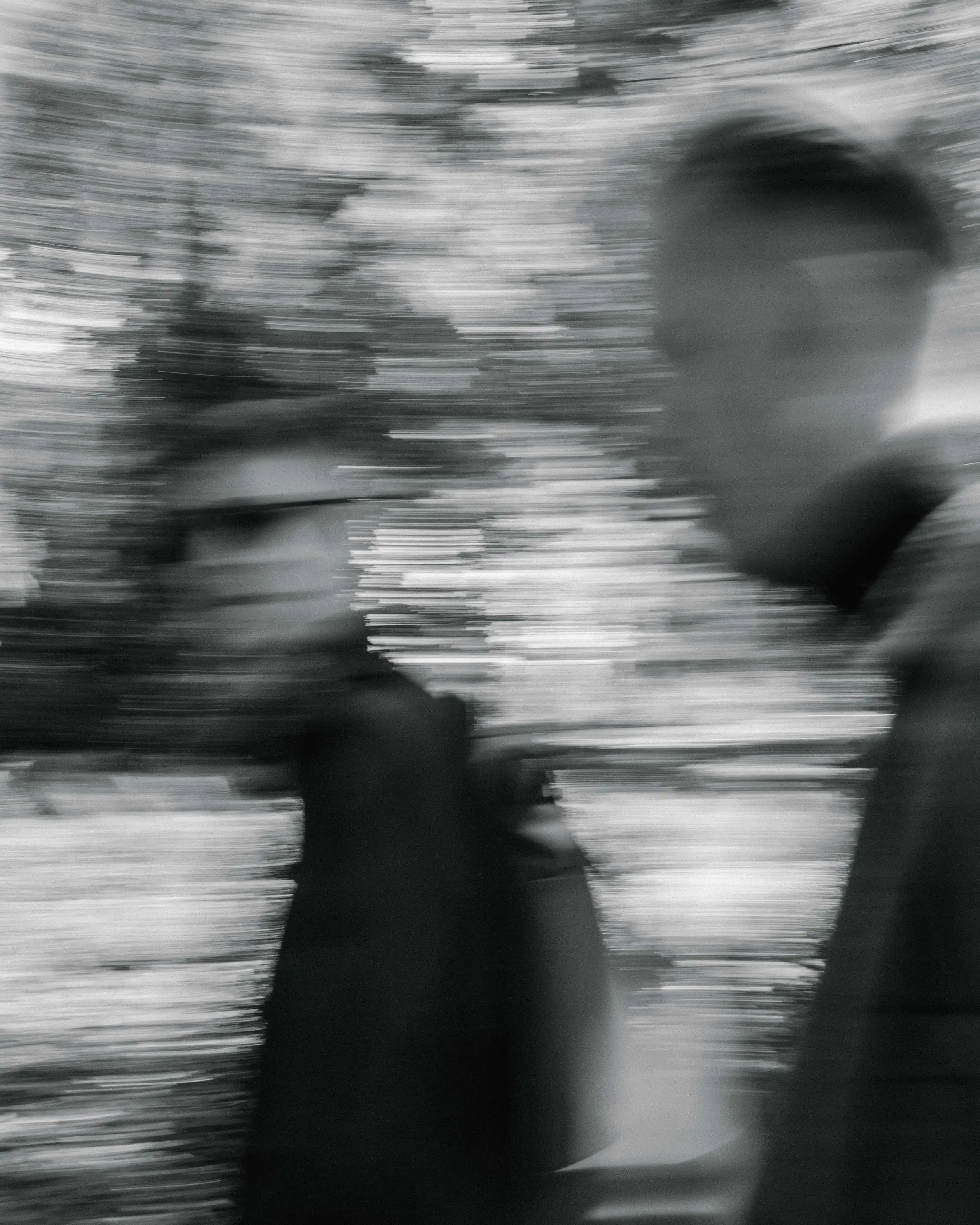 Black-and-white motion-blur photo of two people walking, a man's face visible in the foreground with a second figure in a cap behind him.