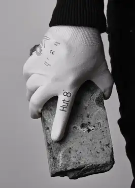 A close-up photo of a person's hand wearing a white Hut 8 branded work glove, gripping a rough gray concrete block or brick against a plain light gray background. The person wears a black long-sleeve top and dark trousers. The glove is printed with the "Hut 8" wordmark along the index finger and small technical markings on the back of the hand — including the Hut 8 sunburst logo, a "CE" safety mark, a size, and cut-level specifications — giving it an industrial, safety-equipment look.