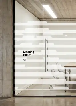 A glass-walled meeting room in a modern industrial office with exposed concrete walls, ceiling, and polished floor. The glass door and partition are covered with a frosted horizontal-stripe privacy film in the Hut 8 brand style, through which a conference table and wire-frame chairs are visible inside. Black text on the glass reads "Meeting Room" with the room code "A2" below it. A recessed vertical door handle and a linear ceiling light complete the clean, minimalist scene.