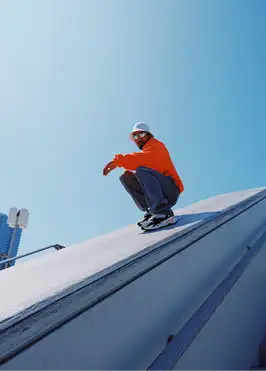 A low-angle shot looking up at a man crouching at the top of a large, pale concrete slope or ramp against a clear blue sky. He wears a bright orange hoodie, dark loose jeans, chunky black-and-orange sneakers, a white beanie, and sunglasses, resting his forearms on his knees as he looks down toward the camera with a relaxed pose. A high-rise building and a streetlight are visible in the lower-left corner. The image has a bold, sunny, streetwear-editorial feel with strong orange-and-blue color contrast.