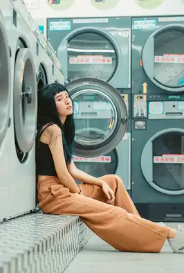 A young woman sits on the floor of a laundromat, leaning back against a row of front-loading washing machines. She has long dark hair with blunt bangs and looks directly at the camera with a calm expression. She wears a black sleeveless crop top and wide-legged tan trousers with white sneakers, one knee drawn up and an arm resting on it. Behind her, stacked teal industrial washers and dryers (branded "Primus") fill the frame, marked with red Chinese-character signage and numbered panels. The scene has a soft, muted, cinematic color palette.