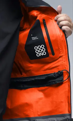 A close-up of a hand holding open the interior pocket of an orange rain jacket, revealing a black fabric patch with a stacked-circle "OOONO" logo and the text "Raincoat 10.000 mm."