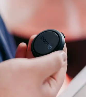 An extreme close-up of a hand holding a small black circular "OOONO" device, against a softly blurred warm orange background.